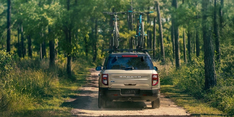 A tan 2024 Ford Bronco Sport driving down a dirt trail on a sunny day with two bikes secured to the vehicle's roof rack