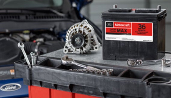Ford Motorcraft vehicle battery alongside an alternator with an open car hood in the background