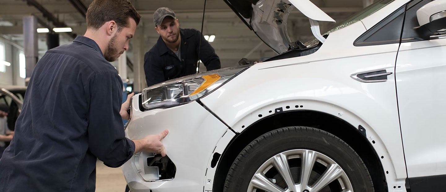 Two mechanics with open hood, one fitting a bumper onto a white car, the other watching closely.