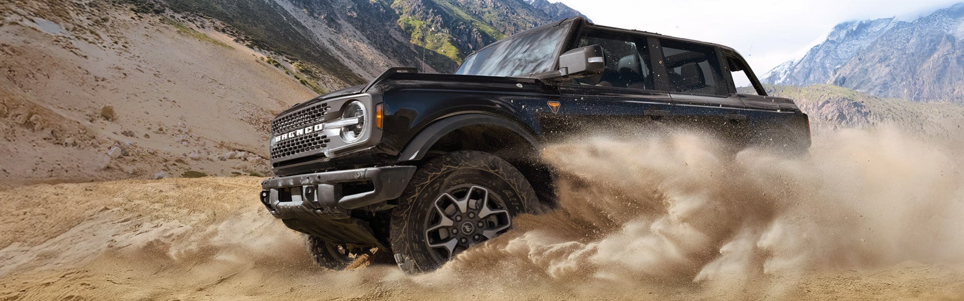A black Ford Bronco four-door SUV kicking up a large cloud of sand as it drives through a desert landscape.