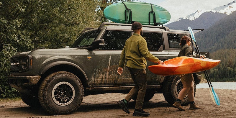 A muddy Ford Bronco with two people carrying kayaks and paddles, parked by a lake with mountains.