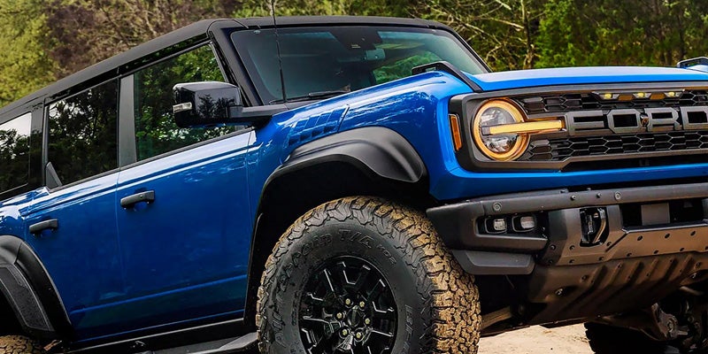 Close-up of a blue Ford Bronco with thick off-road tires, black fender flares and grill, and orange headlights.