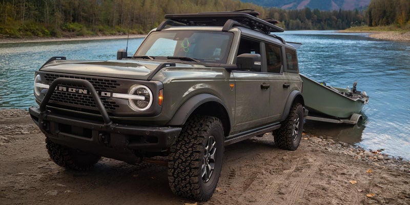 A green Ford Bronco SUV pulls a boat on a trailer to the edge of a blue lake, surrounded by mountains.