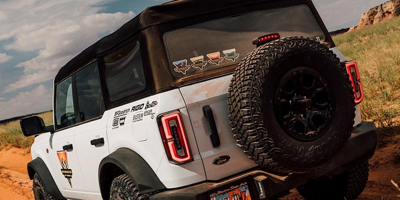 Rear view of a white Ford Bronco with black canvas top and large spare tire on the back, driving on dirt.