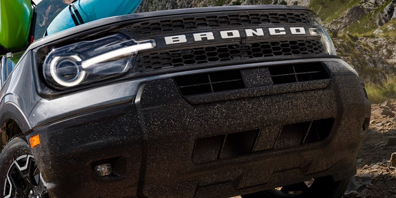 Front view of a dark gray Ford Bronco Sport with glowing headlight, dirty black grille, and two kayaks on top.