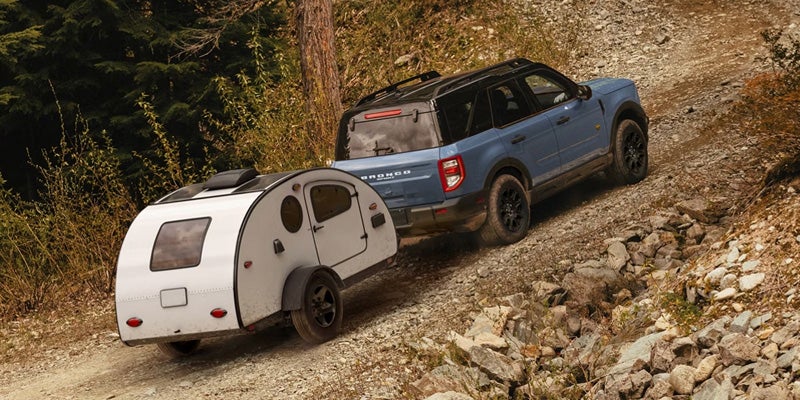 A blue Ford Bronco Sport SUV towing a white teardrop camper downhill on a rocky dirt road in a forest.