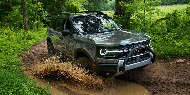 Green Ford Bronco Sport splashing through a muddy rut on an off-road trail in a dense forest.
