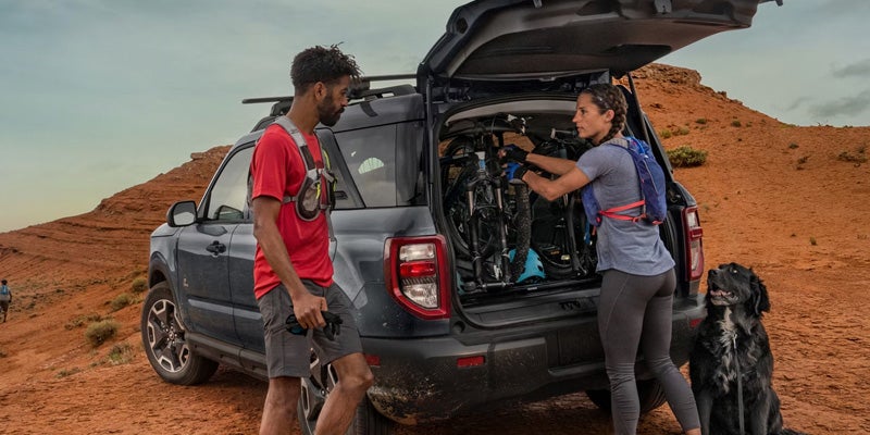 Man and woman load mountain bikes into the trunk of a black SUV in a desert landscape, black dog sitting nearby.