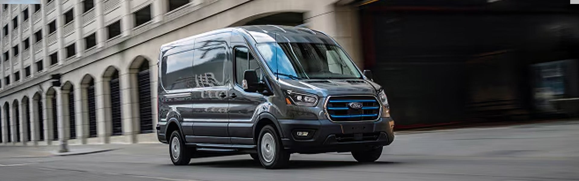 Dark gray Ford E-Transit electric cargo van with blue badging drives down a city street.