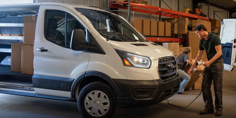 A white electric cargo van with an open side door, being loaded with boxes while charging, in a warehouse.