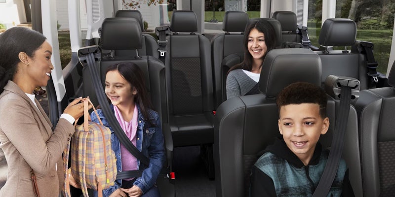 A woman hands a backpack to a smiling young girl seated in a van, with two other children smiling in the back rows.