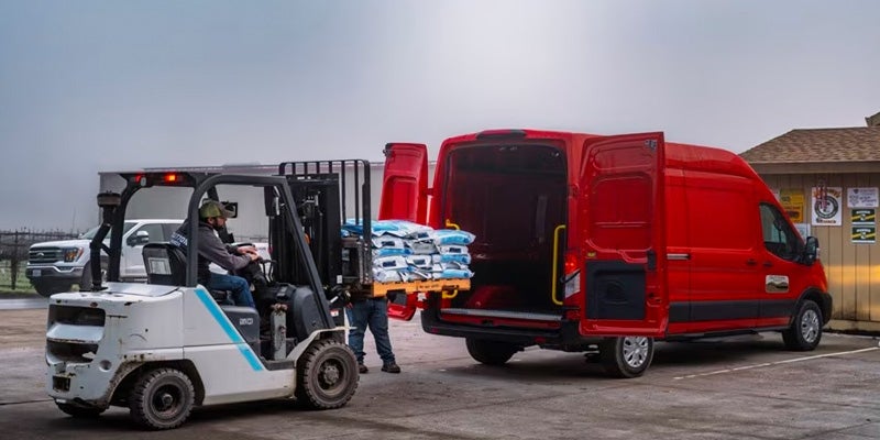 A forklift operator loading bags of product from a pallet into the back of an open red delivery van.