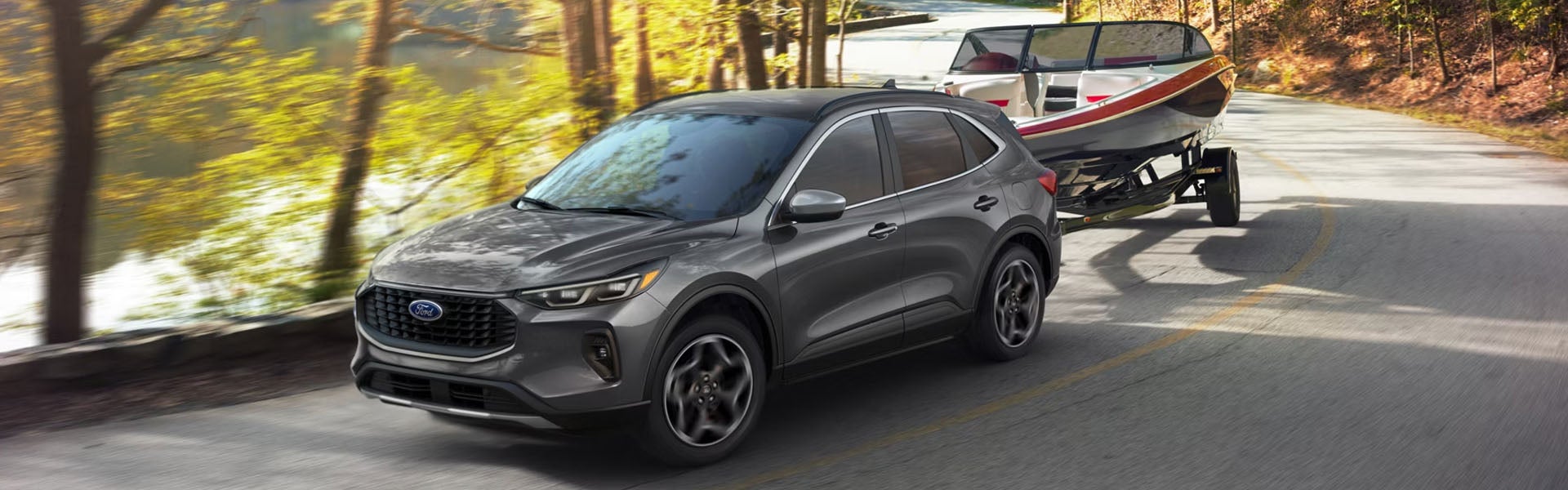 A grey Ford Escape SUV towing a boat on a trailer, driving on a winding road next to a lake and autumn trees.