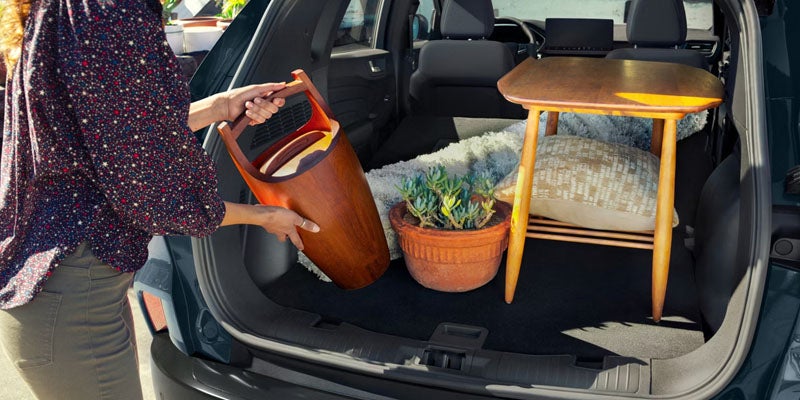 A woman in a floral blouse loads a wooden basket into the trunk of a car already holding a potted plant and a small table.