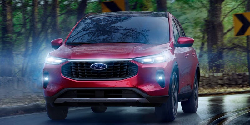 A red Ford SUV with headlights on driving on a wet road lined with trees.