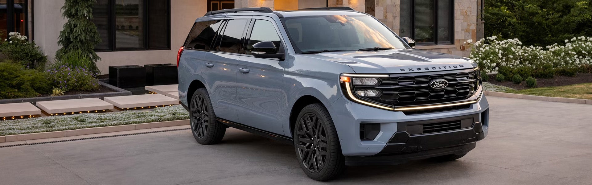 A grey Ford Expedition with black wheels parked on a paved driveway in front of a modern building with landscaping.