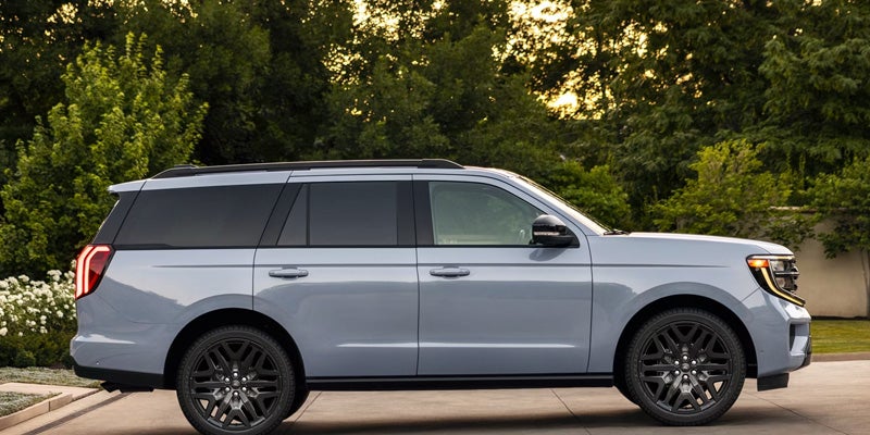 A light blue SUV with black tinted windows and black rims, parked on an asphalt driveway with green trees in the background.