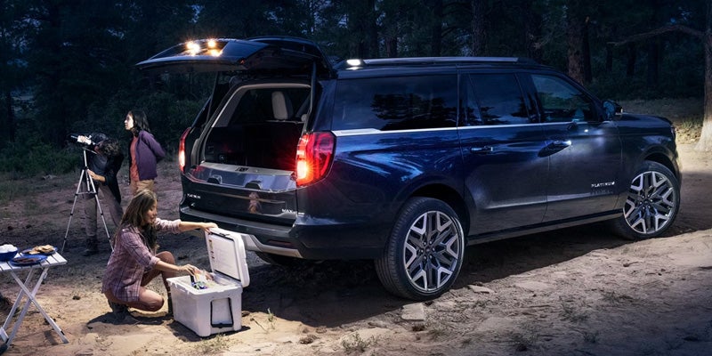 Family camping at night by dark blue SUV with open trunk, using cooler and telescope.