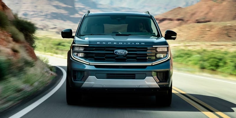 A dark blue Ford Expedition SUV driving head-on down a winding road with rocky hills in the background.