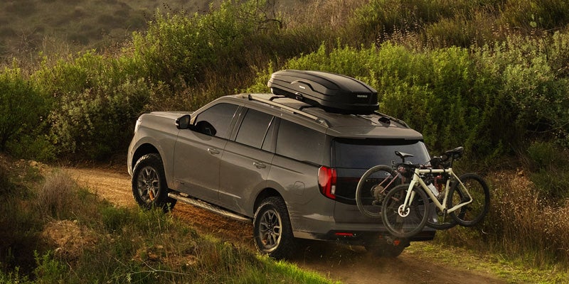 Rear view of a grey Ford Expedition SUV with a roof cargo box and two bikes on a rear rack, driving off-road.