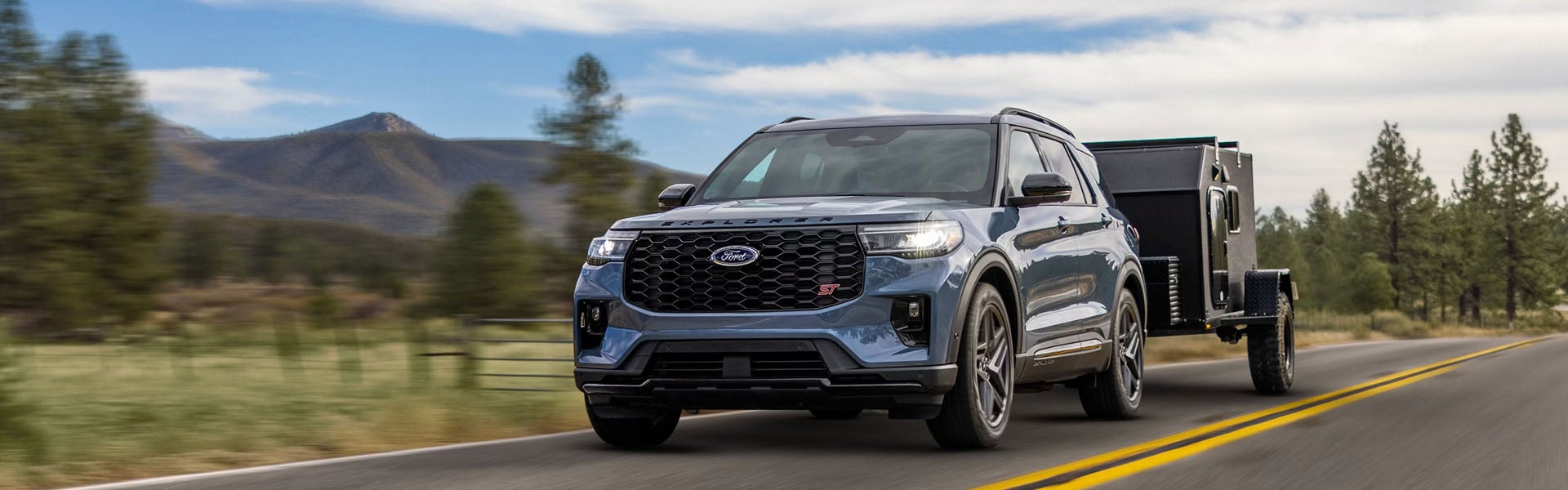 A blue Ford Explorer ST SUV towing a black teardrop camper on a mountain road with trees and a cloudy sky.