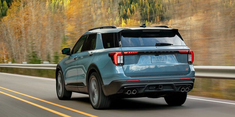 Rear view of a driving Teal Ford Explorer ST on a road with autumn trees in the background.