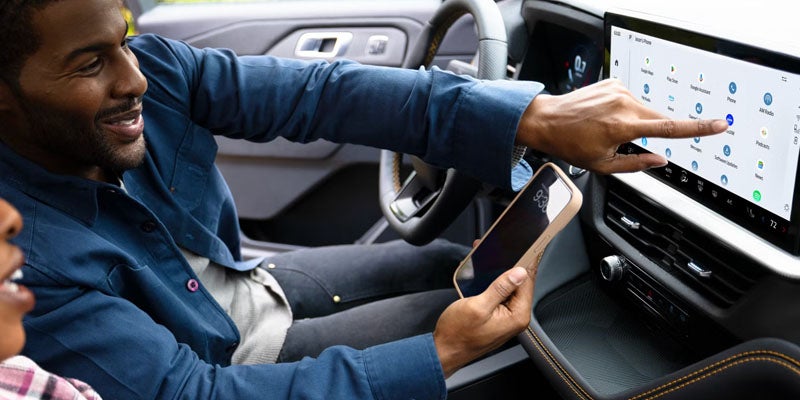 A smiling man in a car, holding a phone in one hand and touching the car's touchscreen display with the other.