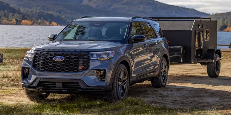 A grey Ford Explorer SUV with a black camping trailer, parked on grass by a lake with mountains in the background.