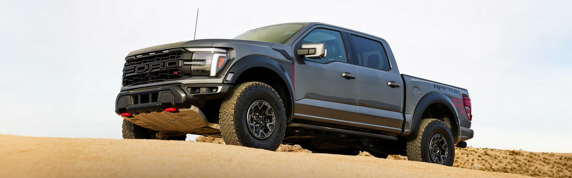 A gray Ford F-150 Raptor pickup truck, with red accents, drives up a sandy dune.