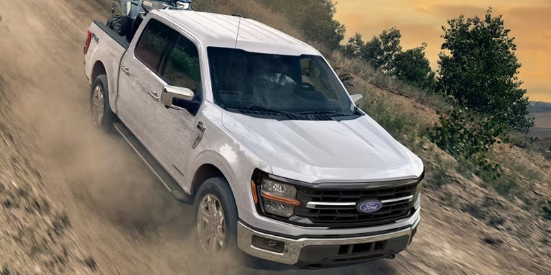 A white Ford F-150 pickup truck with a four-wheeler in its bed drives up a dusty dirt road.