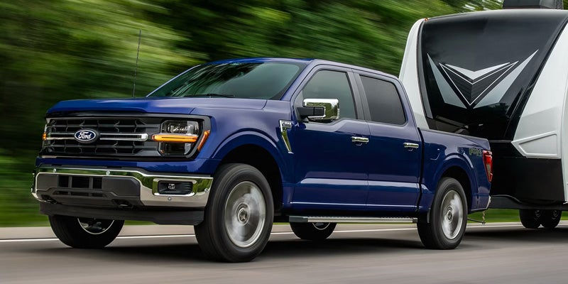 Blue Ford F-150 pickup truck towing a white and black travel trailer on a highway.