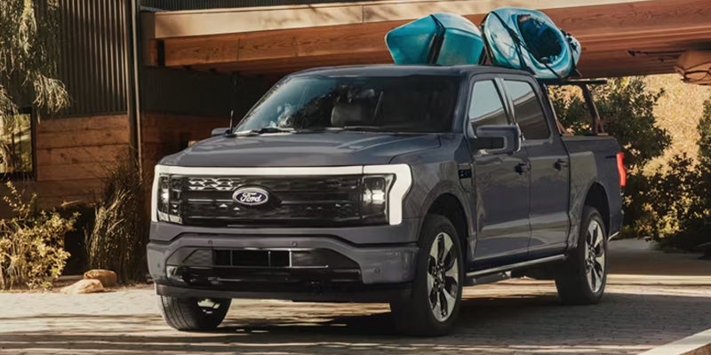 Dark gray Ford F-150 Lightning electric pickup truck, with two blue kayaks on top, parked in front of a rustic building.