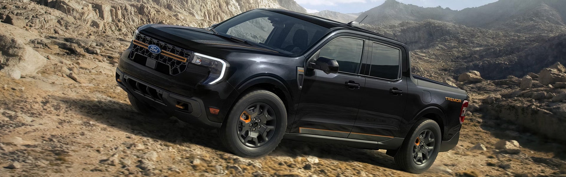 A black Ford Maverick Tremor pickup truck with orange accents drives up a rocky path with a mountain range in the background.