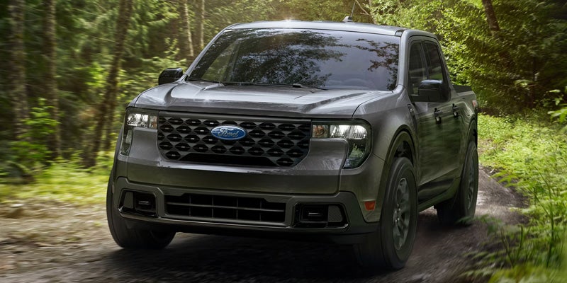 A dark gray Ford Maverick pickup truck drives toward the viewer on a dirt road through a sun-dappled forest.