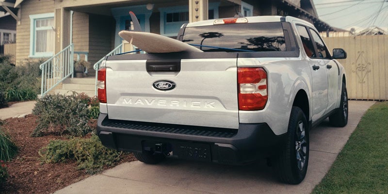 Rear view of a Ford Maverick pickup truck with a surfboard in its bed, parked in a driveway.
