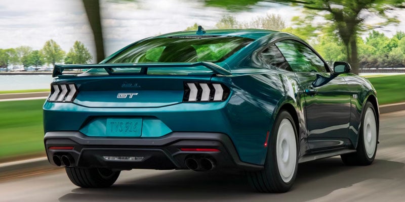 Rear view of a teal Ford Mustang GT driving on a road, with a body-color spoiler, dual exhausts, and distinctive taillights.