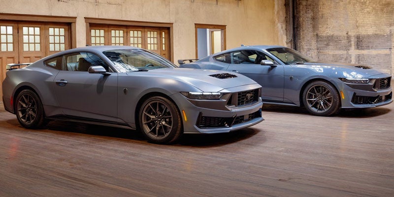 Two new grey 2024 Ford Mustang Dark Horse coupes are shown from the side in a showroom with wooded walls.