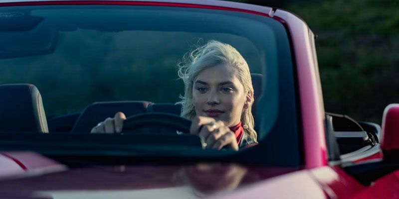 A young blonde woman in a red shirt with windblown hair, driving a red convertible with her hands on the wheel.