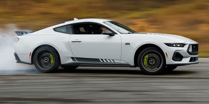 Side profile of a white 2024 Ford Mustang GT drifting on an asphalt track, smoke billowing from its rear tires.
