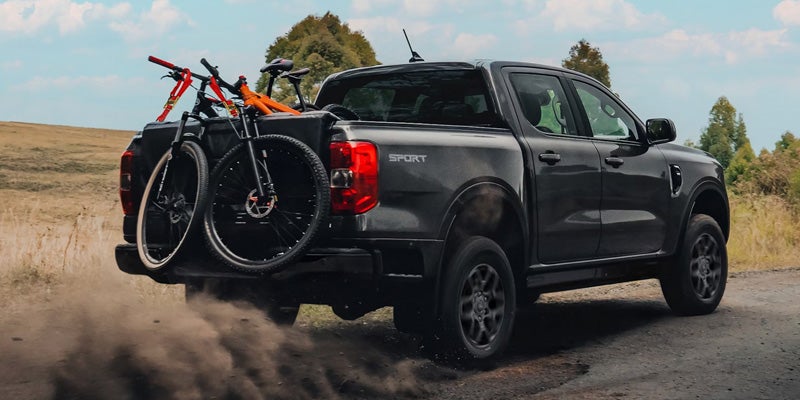 Dark gray pickup truck with two bicycles in the bed, driving on a dirt road, kicking up dust.