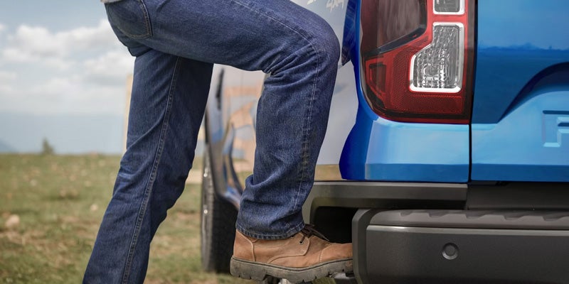 A person in jeans and work boots steps onto the integrated bumper step of a blue pickup truck.