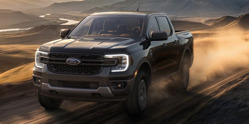 Black Ford Ranger pickup truck kicks up dust on a dirt road in a desert landscape with mountains and a river.