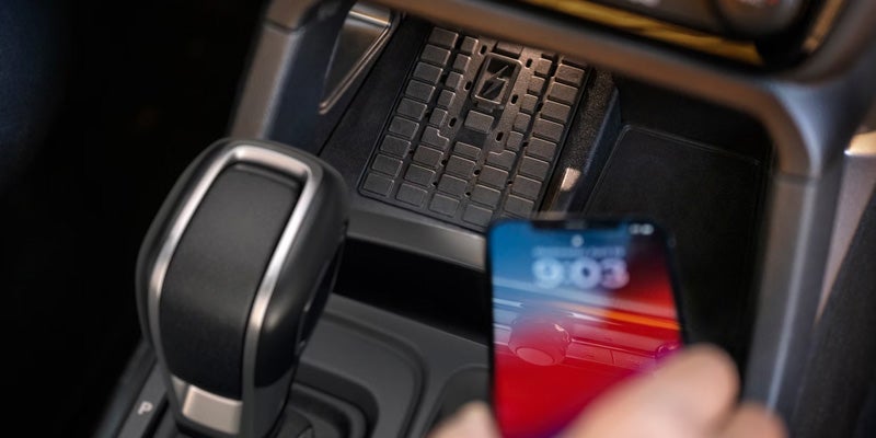 A person's hand places a smartphone on a black, textured phone holder in a car's center console next to a gear shifter.