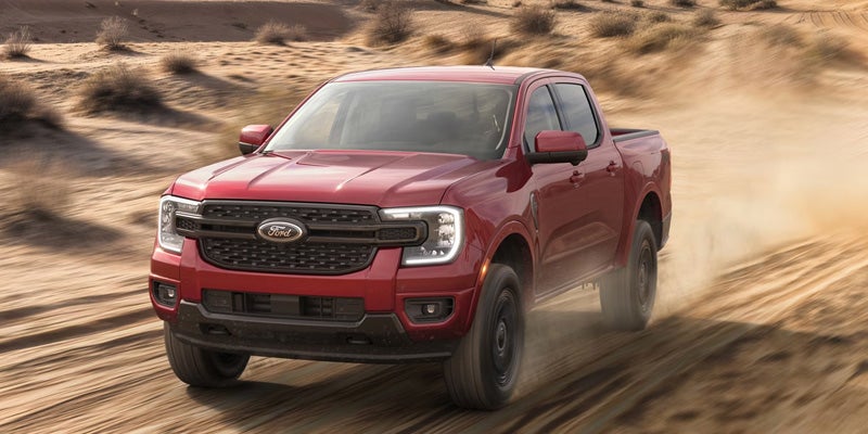 A red Ford Ranger pickup truck kicks up dust as it drives forward on desert dunes with sparse scrub brush.