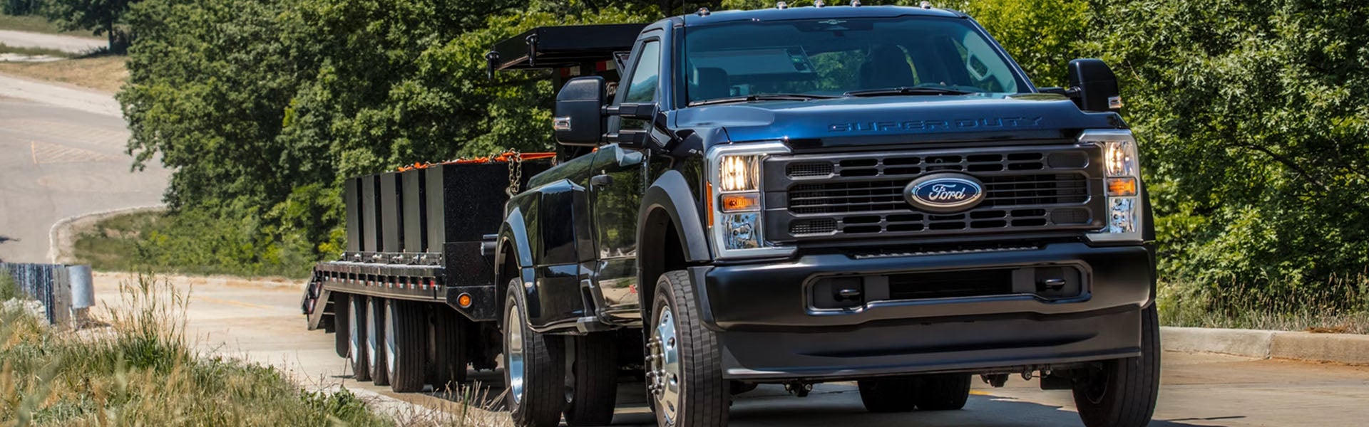 Dark blue Ford Super Duty truck pulling a loaded flatbed trailer on a winding road with green trees and brush.