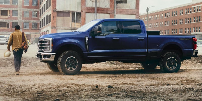 A navy blue Ford F-250 pickup truck with "TREMOR" on the bed, parked on a dirt lot, construction worker walks away.