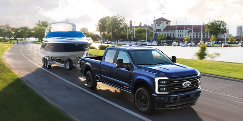 Blue Ford F-250 pickup truck towing a boat on a trailer, driving on a road beside a marina.