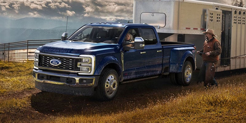 A blue Ford Super Duty pickup truck with a woman driving, towing a horse trailer, with a man standing by.
