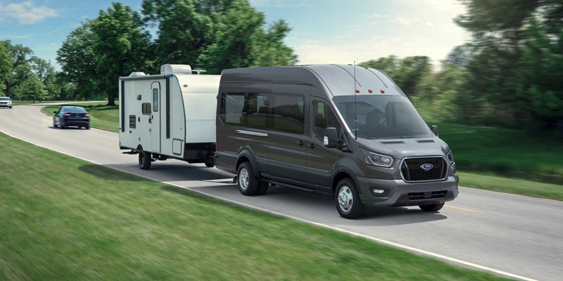 A dark gray Ford Transit van with a white travel trailer driving along a road lined with trees and green grass.