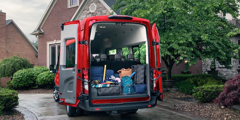 Rear view of a red passenger van with its back doors open, loaded with suitcases and bags, parked in a driveway.